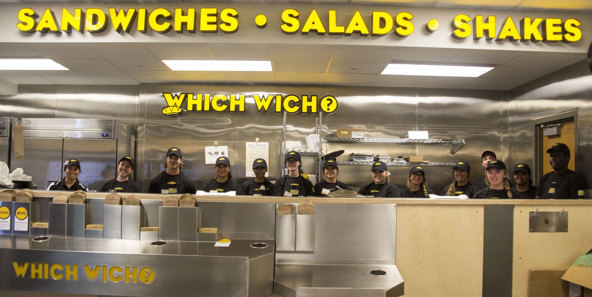 Young adults are lined up behind the counter of the sandwich shop Which Wich.