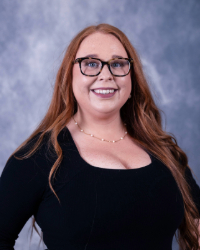 07112025_Amy_C_Related_Image A woman with red hair and black glasses wears a black blouse and smiles in front of a grey backdrop in a professional headshot.