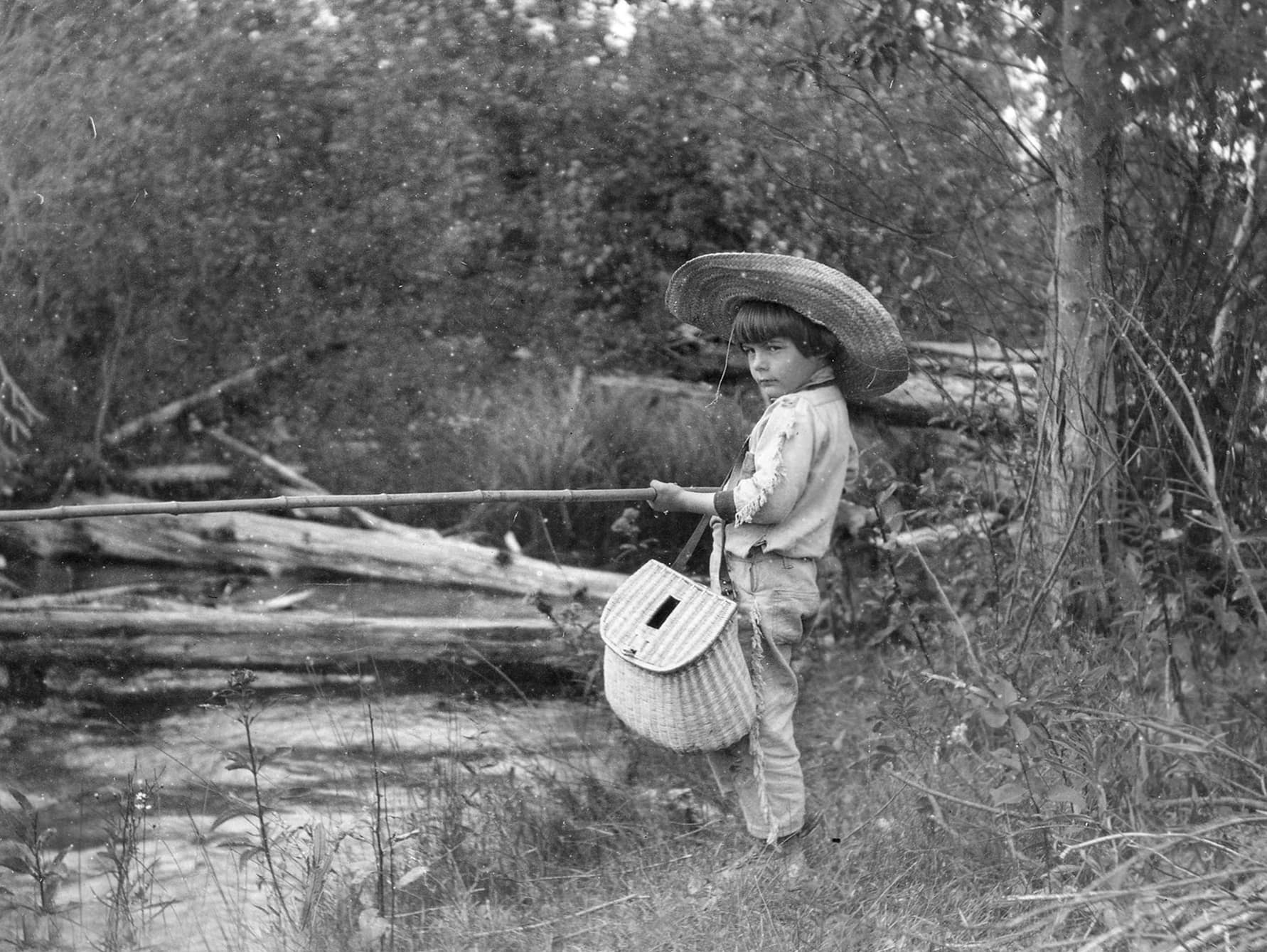 20210723_Support_Hemingway_006 Hemingway as a young boy fishing next to a creek, dressed in large straw hat, a long sleeve shirt, long pants and holding a woven straw bag
