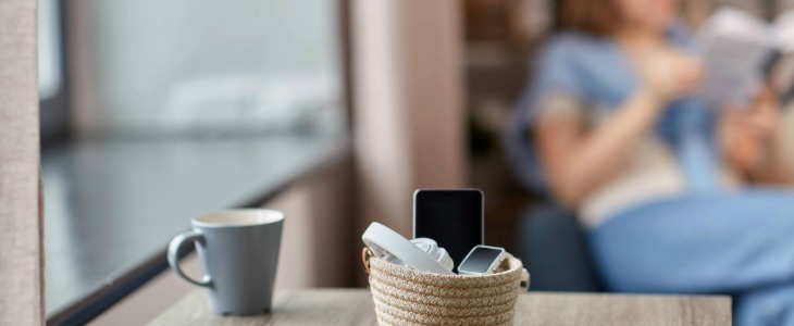 DigDetox-1-Blog_Body Header_730x300 Basket of electronics on a table next to a mug with a person across the room In the background reading a book.