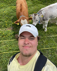 Headshot of Joseph Dinkel in a yellow shirt, white baseball cap, black backpack against a green grass pasture background with one brown and one white gray cow.