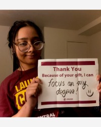 a smiling woman holding a sign that says, Thank you. Because of your gift, I can focus on my degree