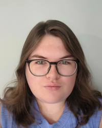 NCharbonneau_maa_headshot Large A woman with brown hair and black glasses wears a blue blouse and smiles in front of a grey backdrop in selfie type headshot.