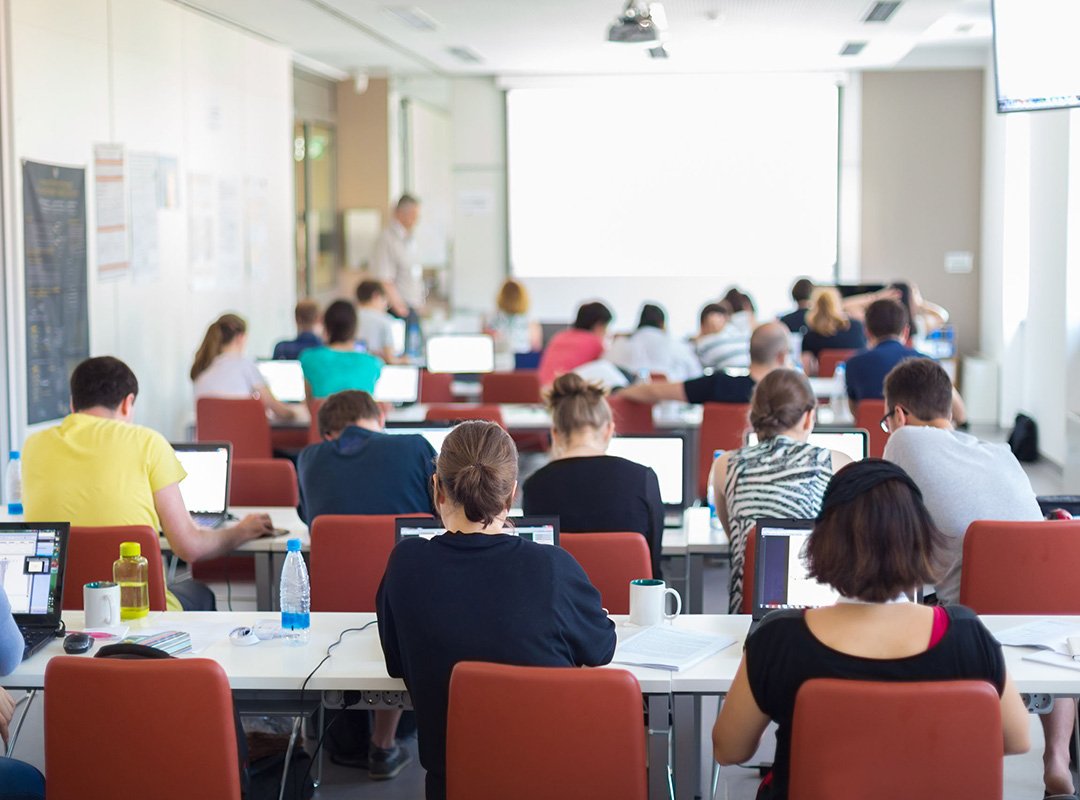 People in a training or classroom working at tables while working on laptops with an instructor.
