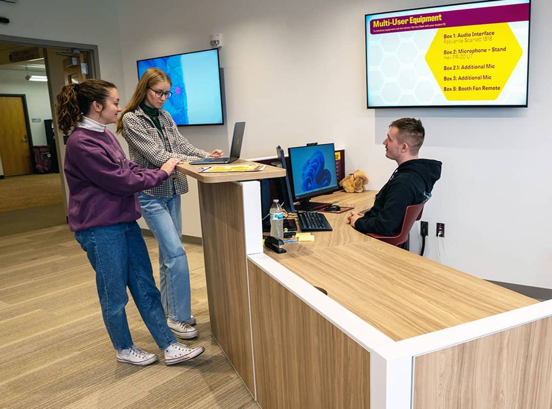 Two students looking at a laptop and asking questions to an Adobe Support student seated behind the desk in the Adobe Digital Lounge
