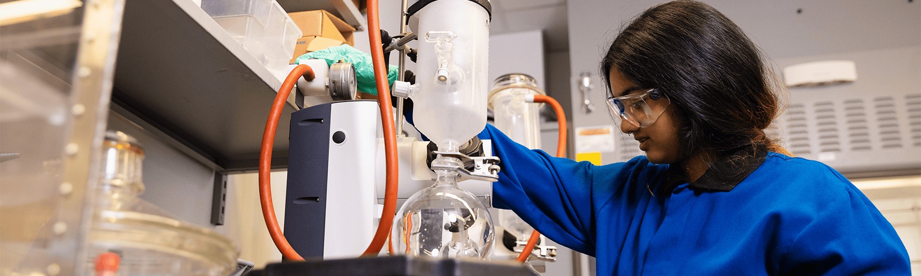 A woman wearing safety glasses and blue lab coat with her hand on a tube looking at a beaker in a laboratory.