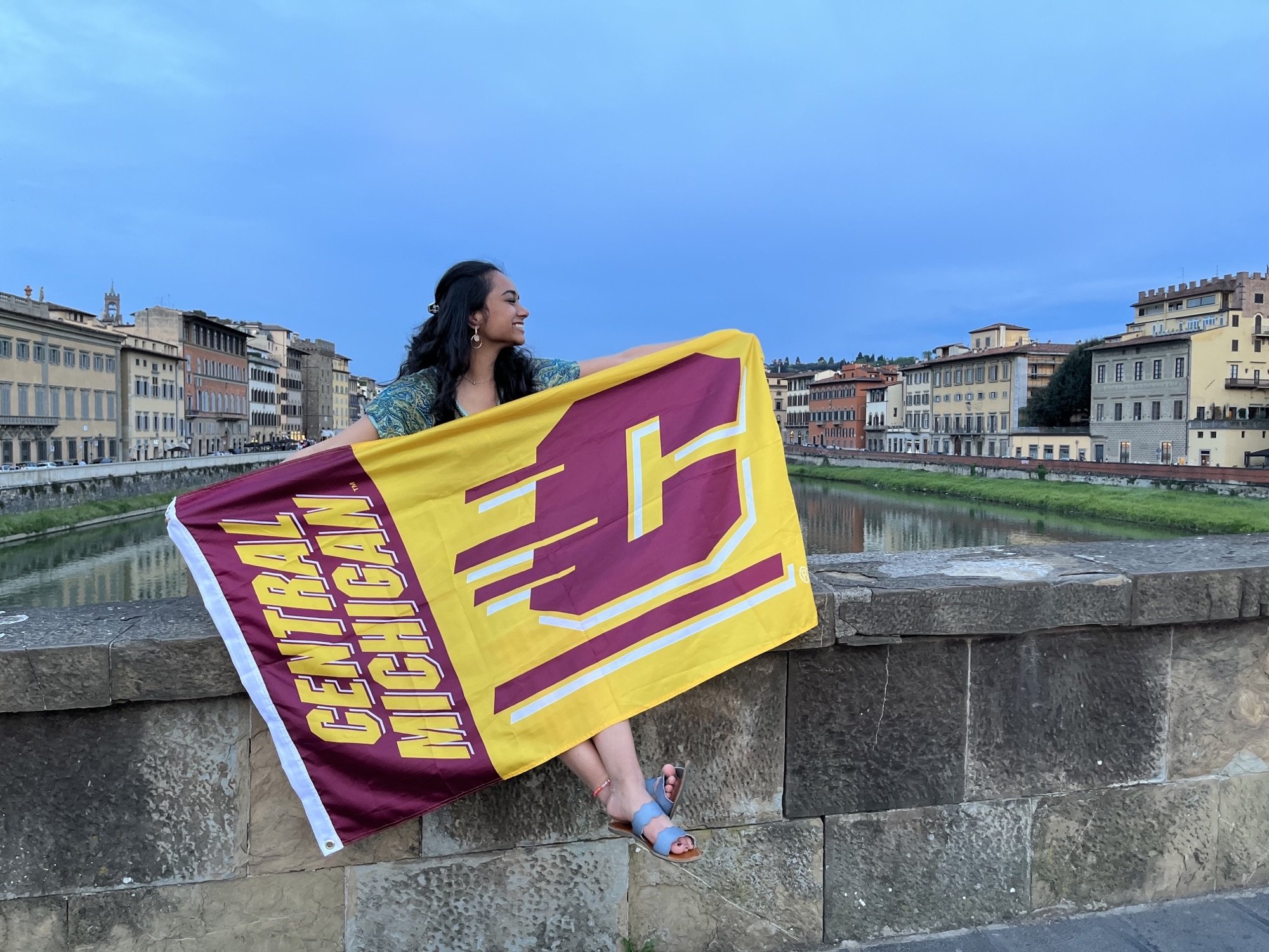 A student sitting on a rock wall holding a Central Michigan University flag.