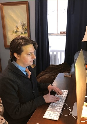 Alexander Rigney wears a suit while sitting at a desk and working on a computer.