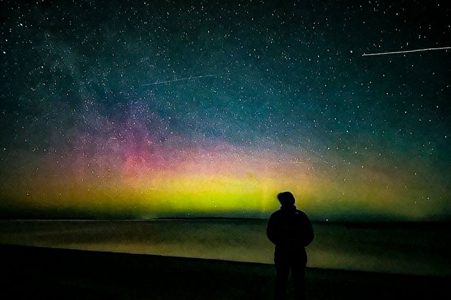 Leah Moskovitz is silhouetted in black against a rainbow display of the northern lights and shooting starts.