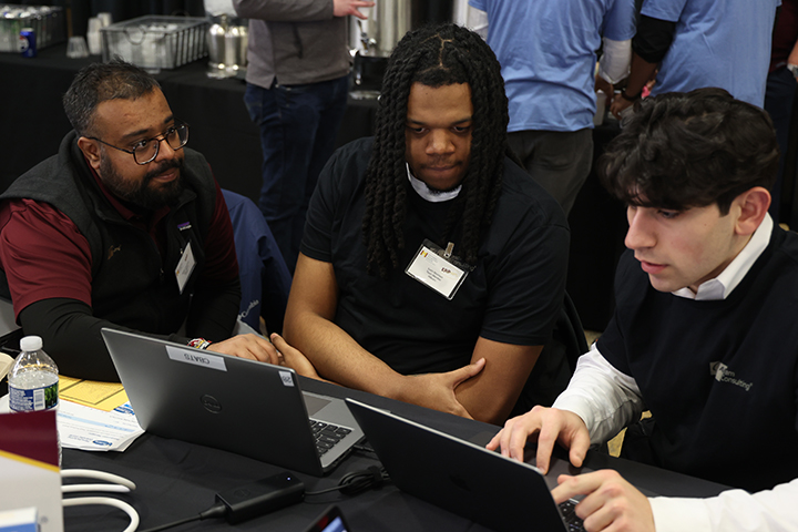 Two students in black t-shirts look intently at a laptop screen. A mentor in a maroon t-shirt and black vest sits to the left. The are in Finch Fieldhouse for the 2025 ERPsim Competition.