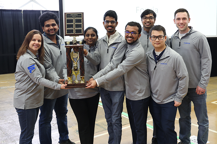 A group of eight students and mentors in longsleeve gray shirts gather around a large trophy and plaque in Finch Fieldhouse for the 2025 ERPsim competition.