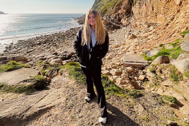 Kennedy Burns stands on a rocky beach in Wales. She's wearing long black pants, a black jacket and gray sweatshirt. She has long blonde hair and smiles at the camera.