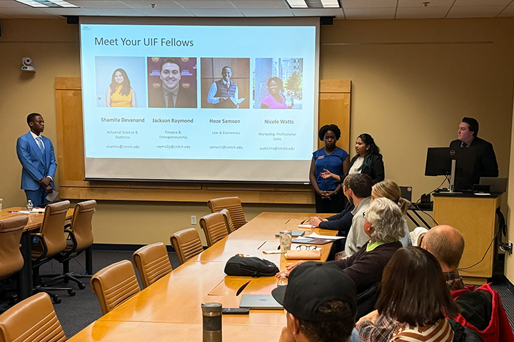 UIF students present to stakeholders in a Park Library conference room. The students stand at the front of the room near a display screen with their bios. Faculty and staff members sit at tables in front of them and listen intently.