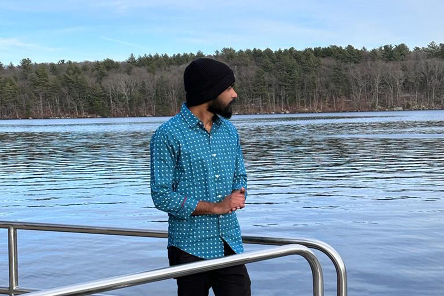 Rambabu Jampana stands on a dock overlooking a lake in the winter. There are evergreen trees in the distance on the shore of the lake. He wears a blue and white spotted button down shirt, black pants and a black winter hat.