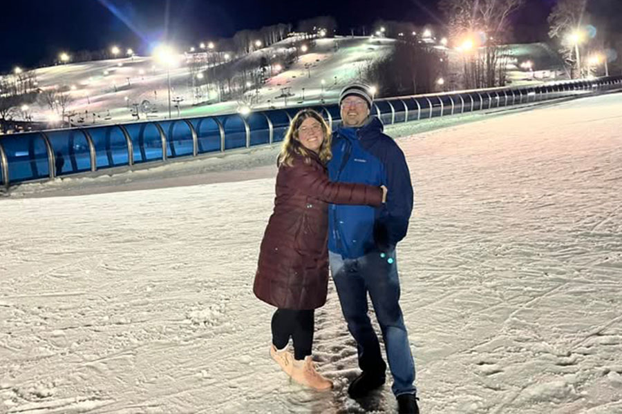 Jasmin Smith stands with her husband in front of a brightly lit ski hill. She's wearing a long maroon coat with black leggings and tan boots. He is wearing black boots, jeans and a blue and navy blue winter coat. 