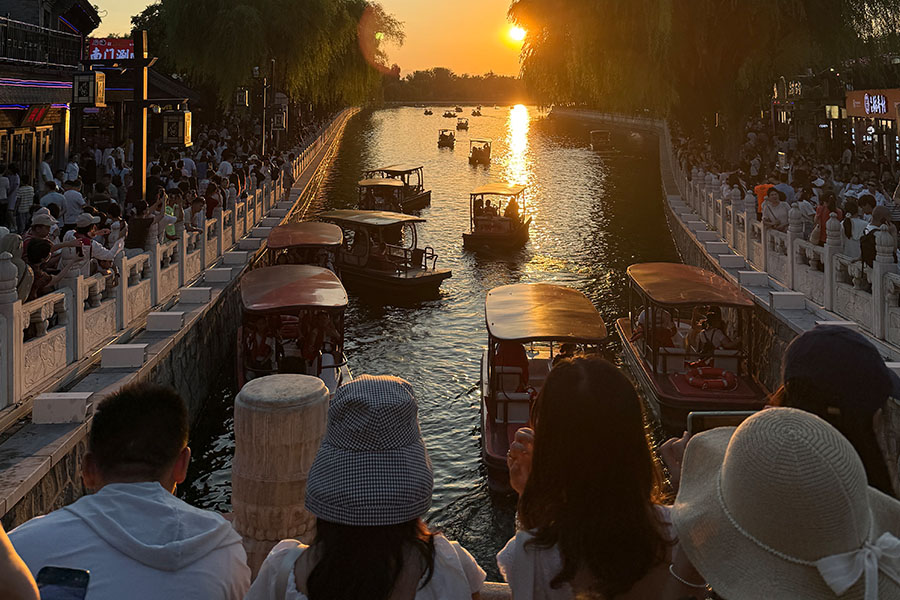 People crowd railings around a Chinese river. They are attempting to capture the sunset and many boats that are on the river. 