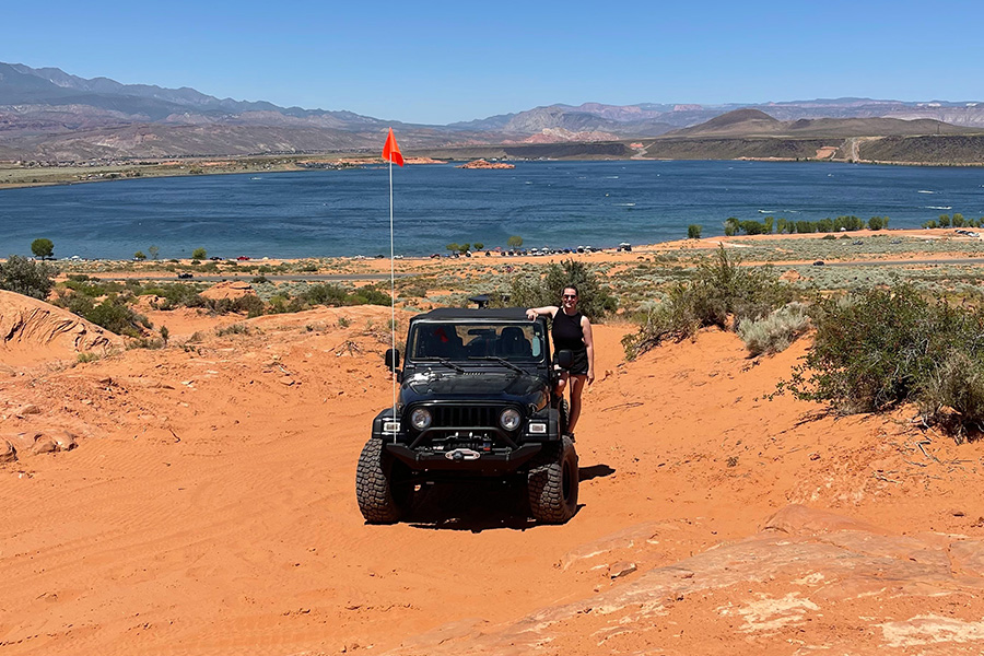 Kelly Potter wears a black tank top and black shorts and stands next to a black jeep with a tall orange flag on a rust-colored sandy overlook. In the distance there is a lake and scrubby brush.