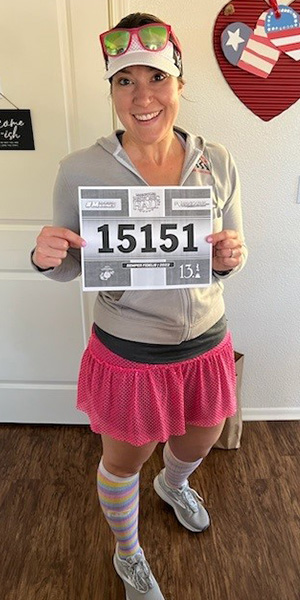 CMU alumna DiAnna Marler poses in her running clothes, which include a hot pink skirt and rainbow socks, while holding a race bib.