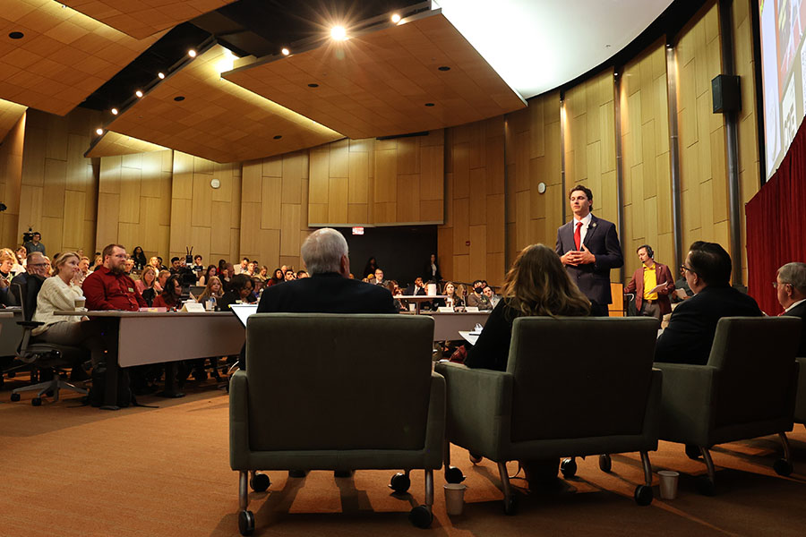 A student in a jacket and tie presents to four judges seated in the front of French Auditorium while a large crowd watches from the seats.