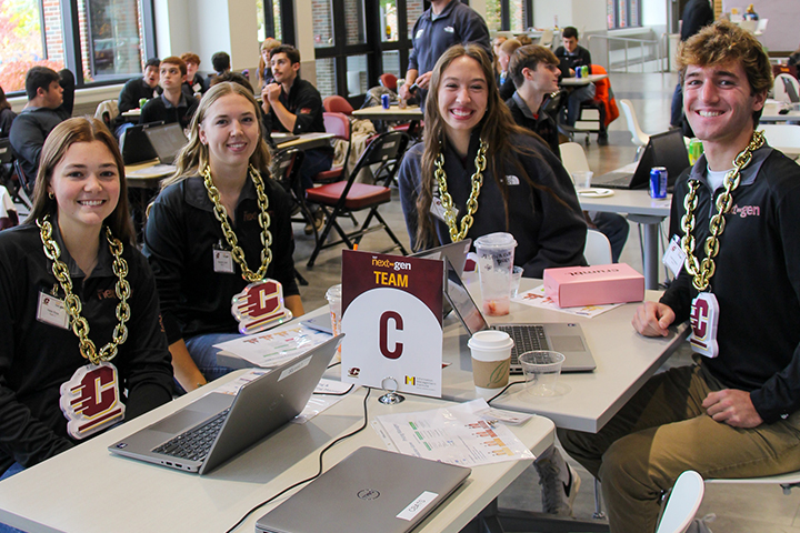 Students in black long-sleeve NextGen shirts wear large plastic gold turnaround chains with action C logos during a high school ERPsim event. The table in front of them has several laptops ready to use for the simulation.
