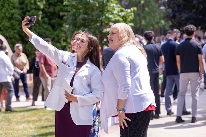 Three women are taking a selfie outdoors during a sunny day. People and greenery are visible in the background.