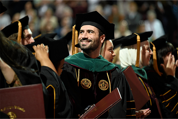 A group of graduates in black gowns and caps stand during a ceremony. One person in the center, smiling broadly, wears a green academic hood and has two medallions on their gown. Others around them also wear gowns and hold diploma folders.