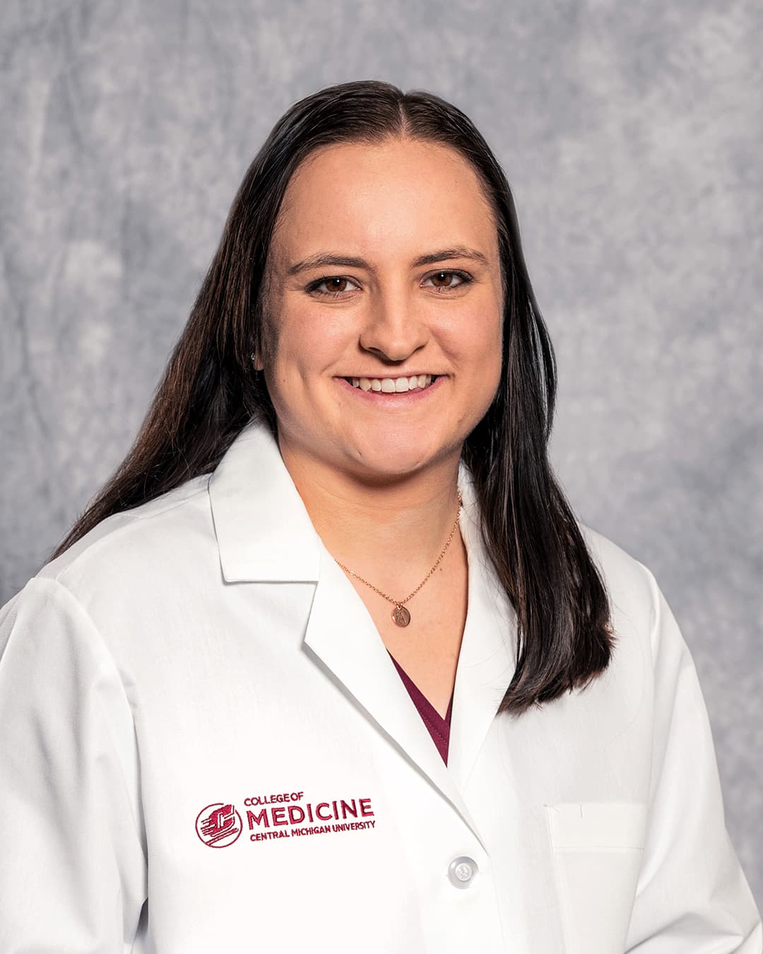 A female CMU College of Medicine medical student with long brown hair wearing her white coat as she smiles for the camera.