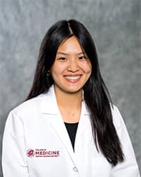 A woman with long, black hair and a black blouse wears a medical white coat and smiles for a headshot.