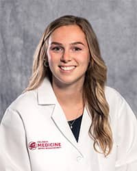 A woman with long, blond hair and a black blouse wears a medical white coat and smiles for a headshot.
