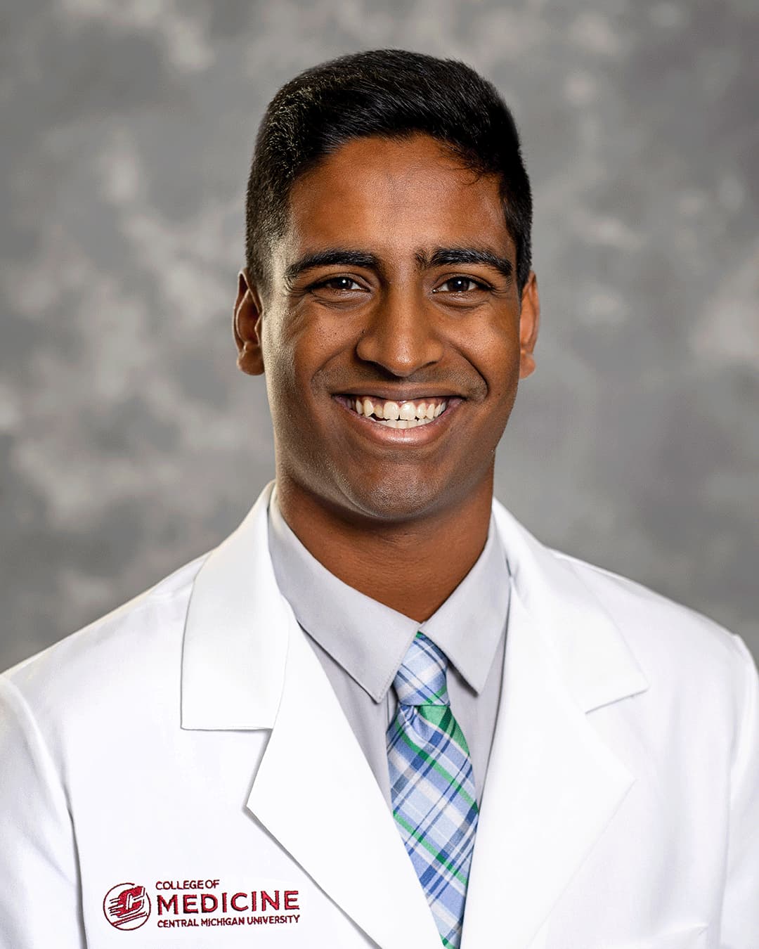 A male CMU College of Medicine medical student wearing a blue and green plaid tie and his white coat as he smiles for the camera.