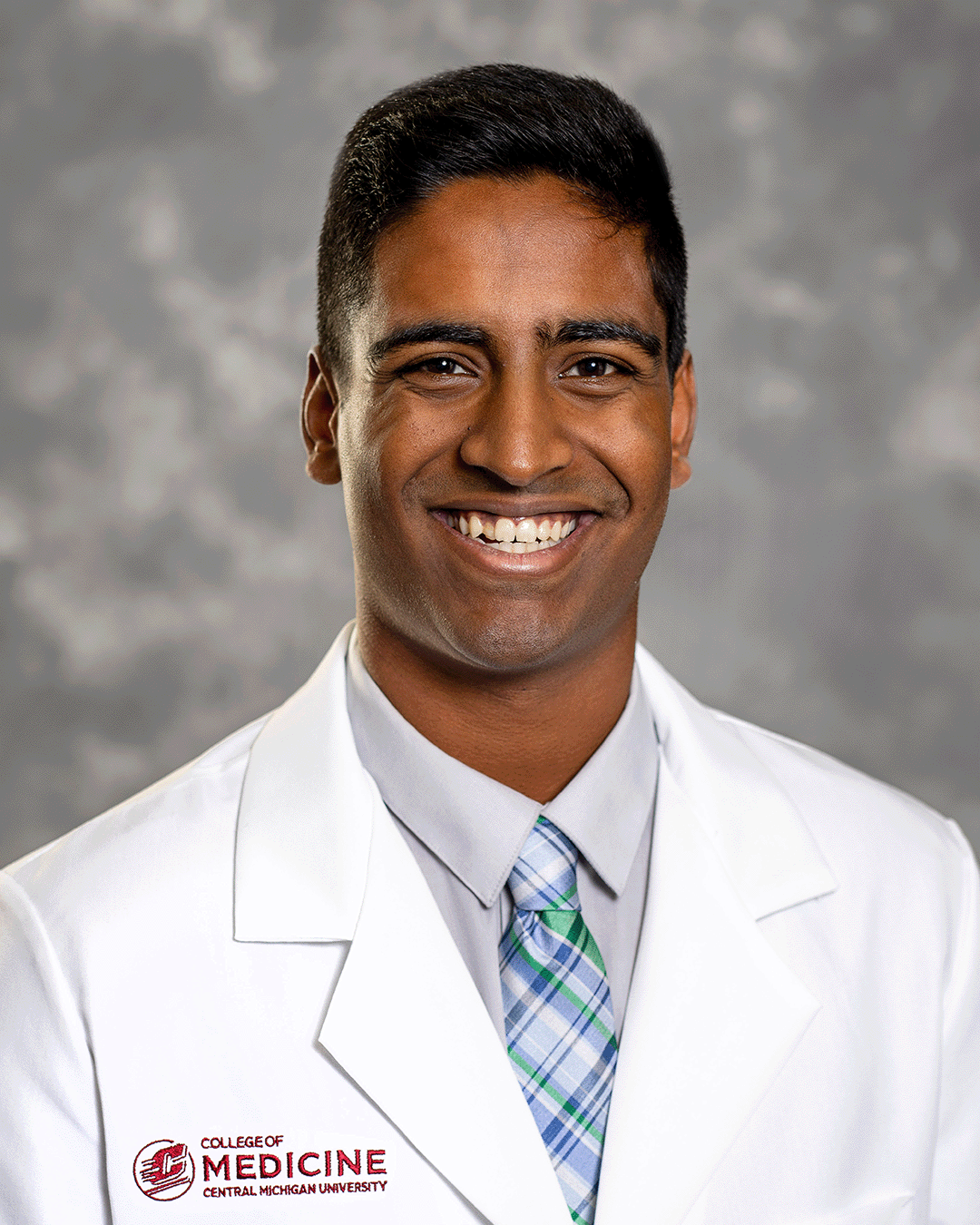 A male CMU College of Medicine medical student wearing a blue and green plaid tie and his white coat as he smiles for the camera.