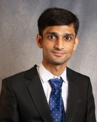 A man with dark hair wearing a dark suit with a white collared shirt and a blue tie.