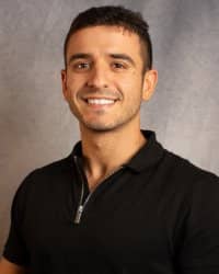 A man in a black shirt smiles for a headshot in front of a grey background.