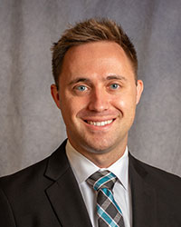 Christopher Prybilla is wearing a black blazer, white shirt, and patterned tie.