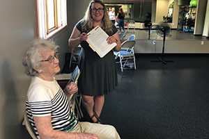 An older woman sits in a chair while a young woman holding a pen and folder smiles.