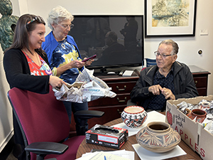 Megan Moreno unwrapping pottery from Jerry Thompson and his wife Mary. The three are gathered around a table that is scattered with more pottery.