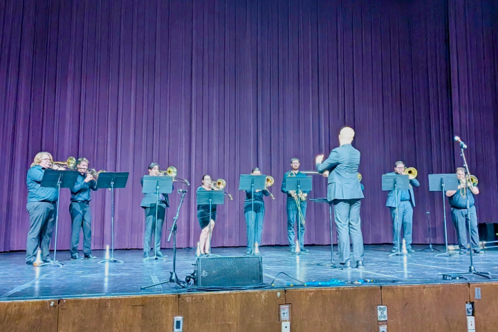 a group of ten people dressed in concert black stand in an arc behind music stands and play trombones on a stage with purple curtains in the background