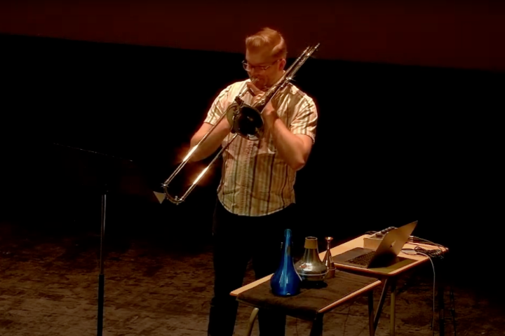 Dr. Cole Bartels playing trombone with mutes and a laptop next to him