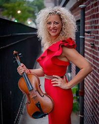 Headshot of Alicia Valoti. She is standing in front of a brick building, holding her viola.