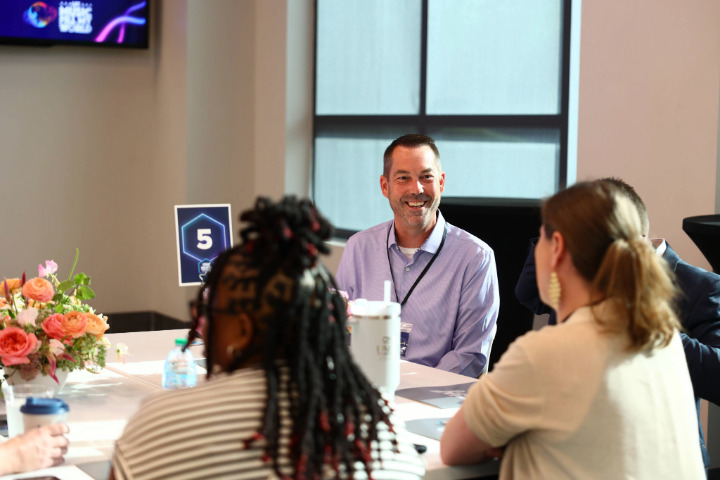 a man smiling at a table with people sitting around it