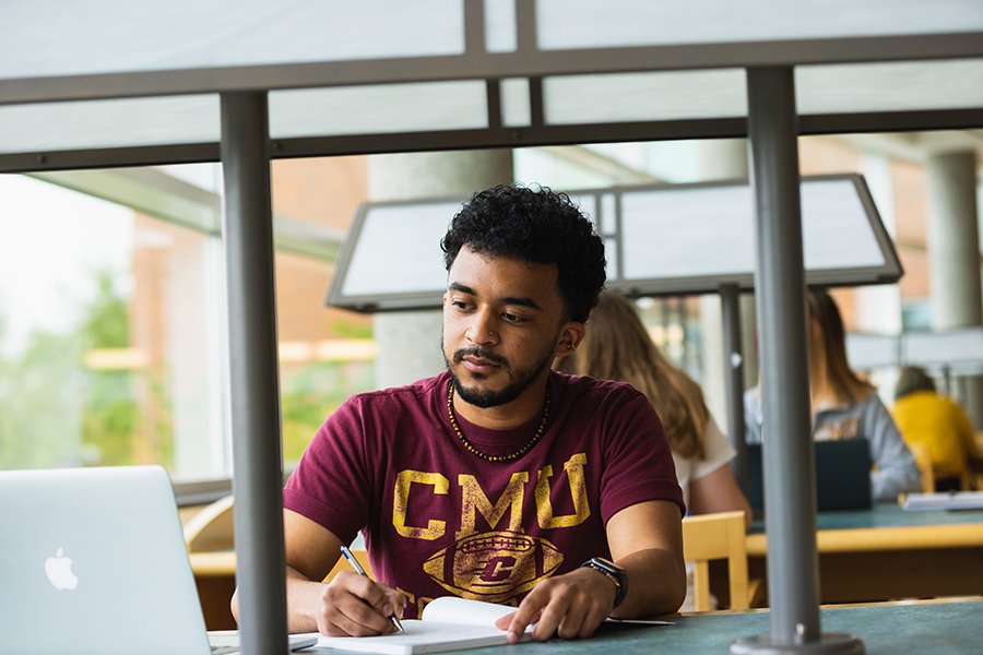 A student sits at a desk with pen in hand, notebook open, and computer open.