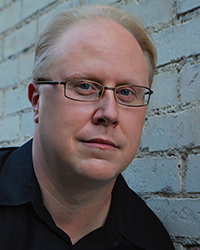 Headshot of Scott Huver in front of a grey brick wall.