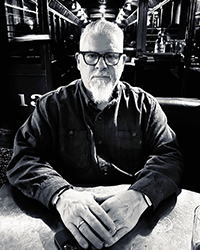 Greyscale headshot of Phil Sgriccia. He's sitting at a table in a restaurant with his hands folded on the table.