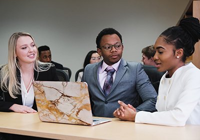 Three students sit at a table in front of a computer, deep in conversation.