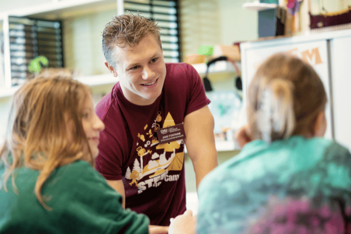 A college student wearing a STEM shirt smiles while listening to two elementary students.