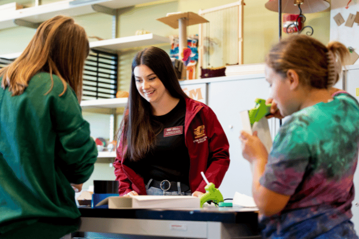 A college student looks on as two middle school students use hot glue guns.