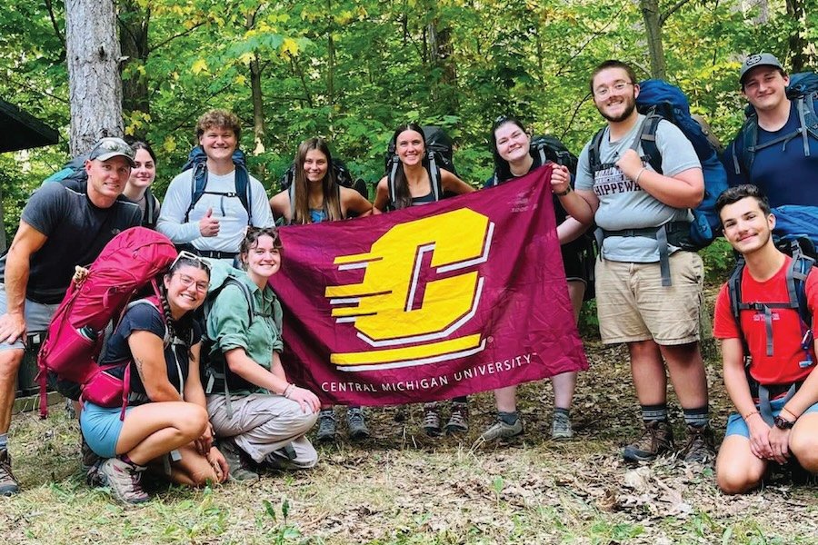 A group of students posing outside in the woods with the large CMU flag.