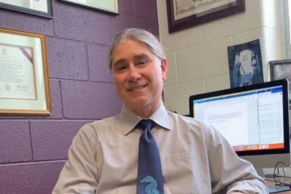 Jeffrey Weinstock, English professor, wearing a collared shirt and blue tie sits at his desk, facing the camera.