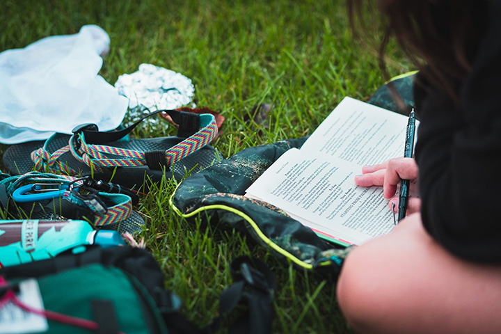 A student sitting on the grass reads from Walt Whitman's Leaves of Grass with a water bottle and shoes on the grass next to the book.