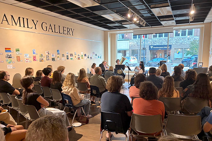 Members of the brass quintet Sassabrass play their instruments while seated in front of large glass windows in front of a room filled with guests seated on chairs.